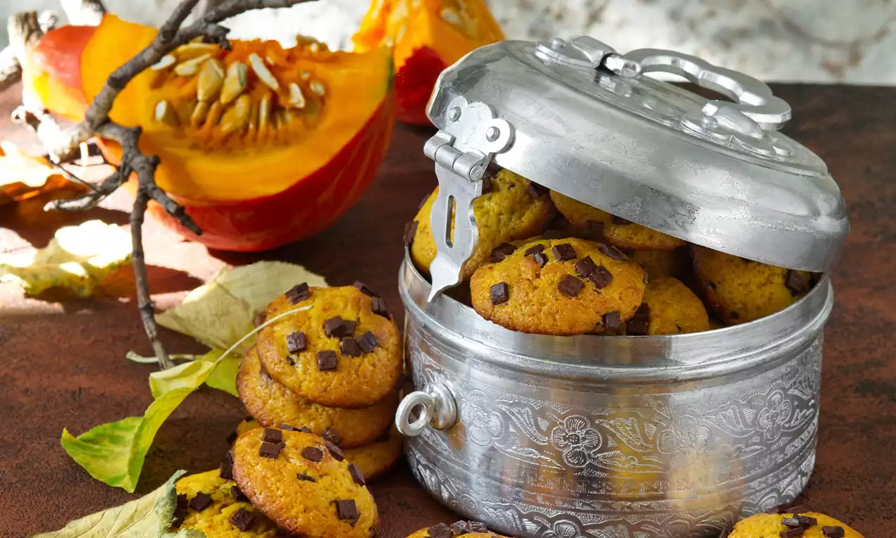 Dýňové sušenky v teplé oranžové barvě s čokoládovými kousky ve stříbrné dóze, pár sušenek vedle a dekorace z dýně a podzimního listí