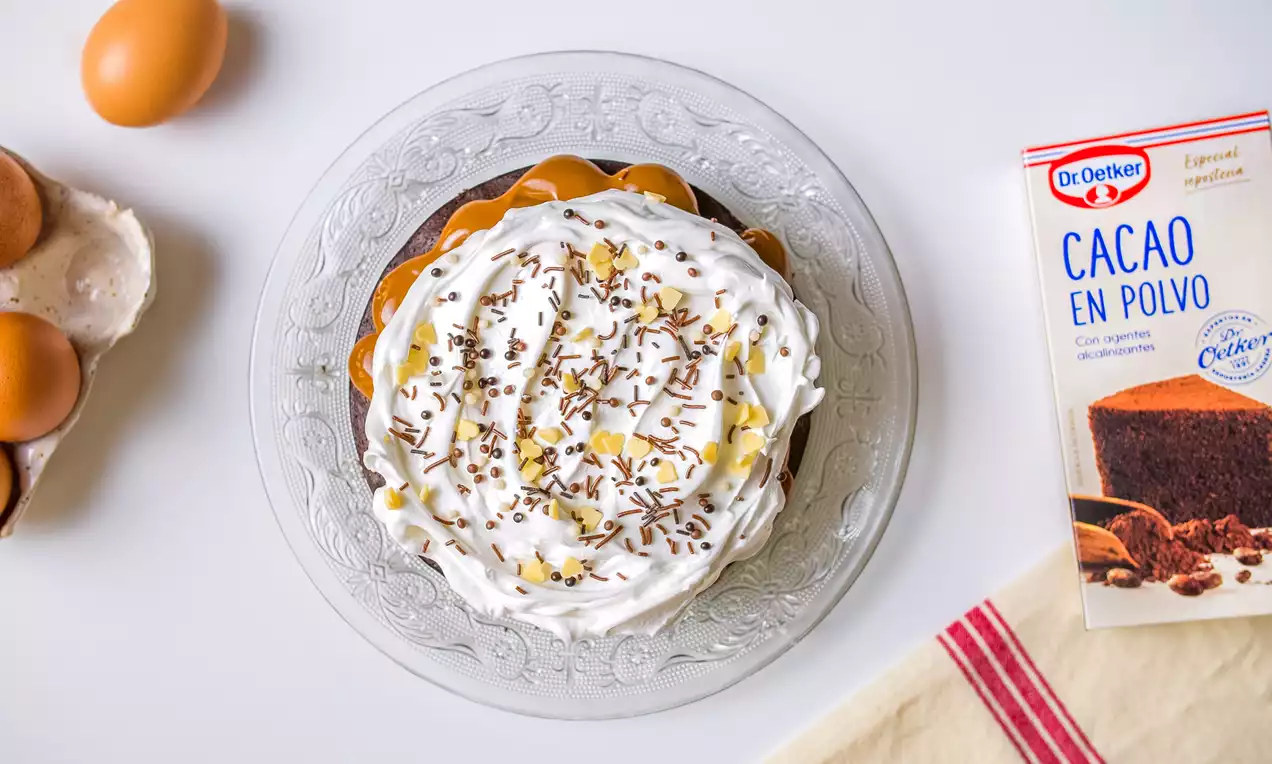 Bizcocho de chocolate oscuro con forma redonda cubierto con crema blanca, trocitos de chocolate y decorado en un plato de vidrio transparente