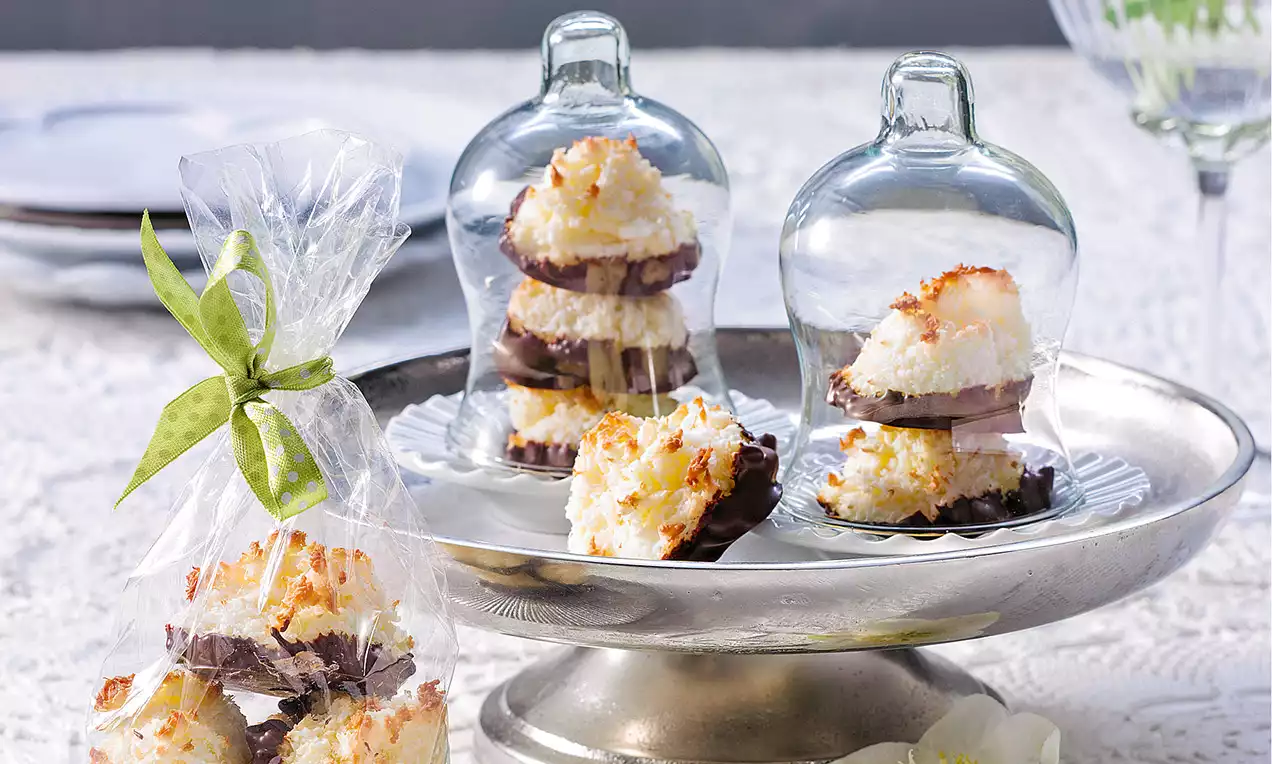 Kokosmakronen mit goldbrauner Kruste und dunkler Schokoladenbasis unter gläsernen Glocken auf einer silbernen Etagere angerichtet