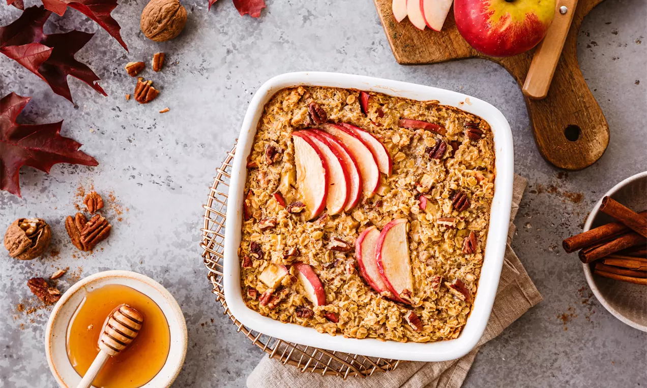 Baked oatmeal rumen-auriu cu felii de măr roșu, nuci pecan crocante și accente de scorțișoară, așezat într-un vas pătrat alb pe un suport metalic
