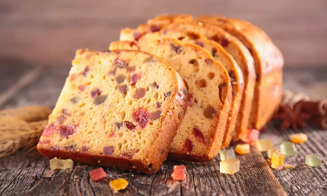 Golden-brown fruit cake slices with colorful candied fruit pieces, arranged on a rustic wooden surface with scattered fruit cubes