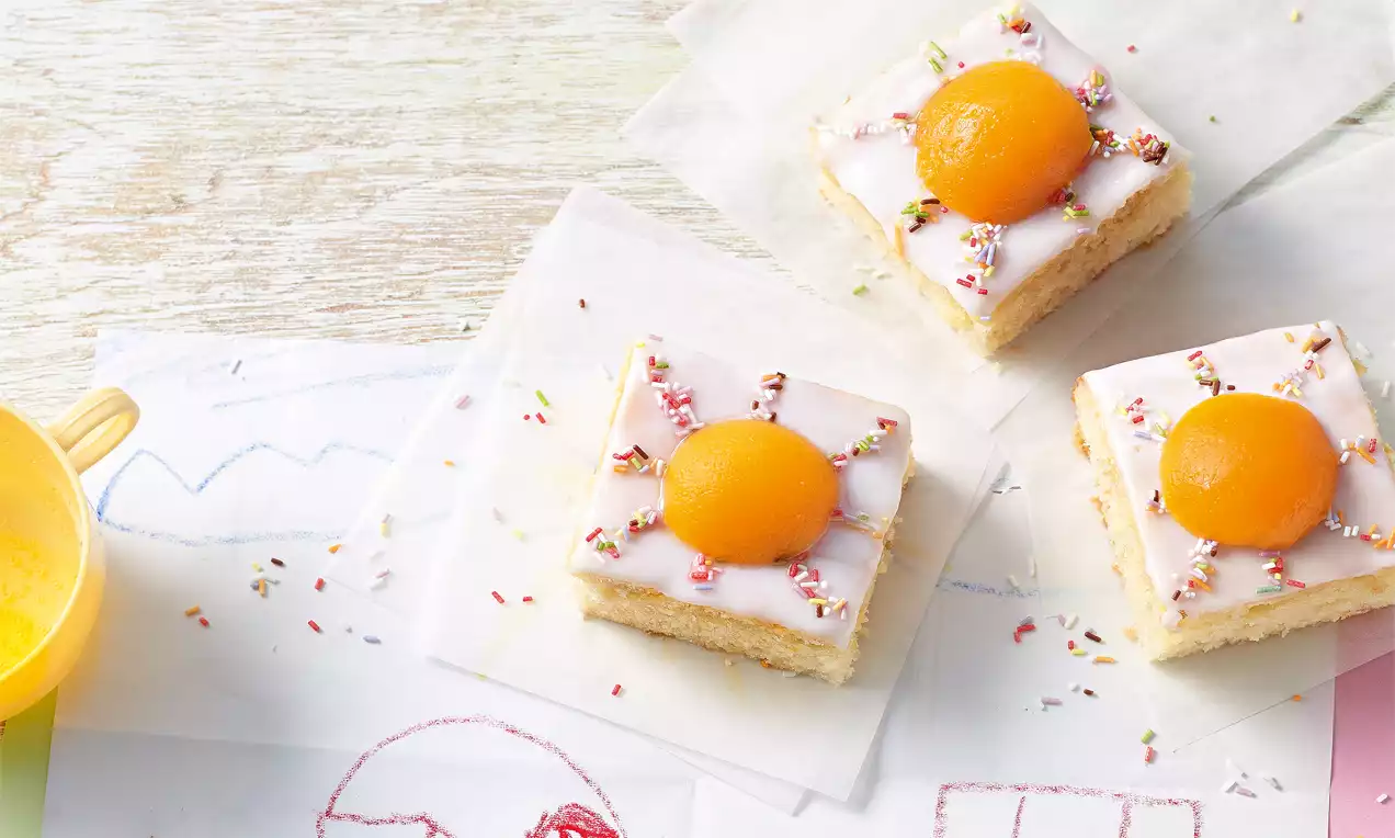 Quadratische Kuchenstücke mit hellem Teig, weißem Zuckerguss, bunter Streusel-Dekoration und einer leuchtend gelben Marillenhälfte auf Backpapier