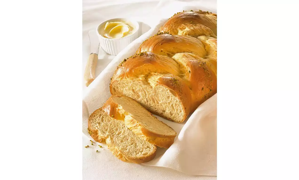 Goudbruine vlecht van luchtig gistdeeg met glanzende korst en enkele plakken op een witte doek, naast een schaaltje boter met mes