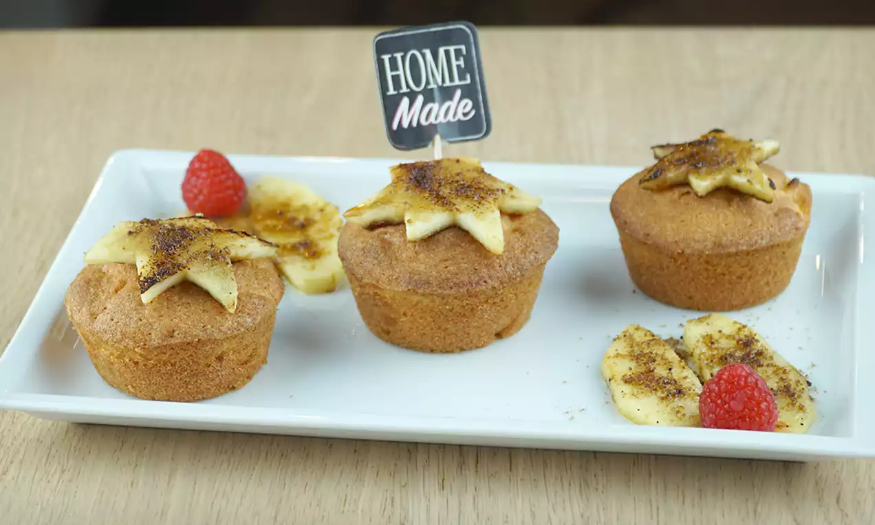Drie goudbruine muffins met gekaramelliseerde bananenschijfjes en frambozen op een rechthoekig wit bord met een houten achtergrond