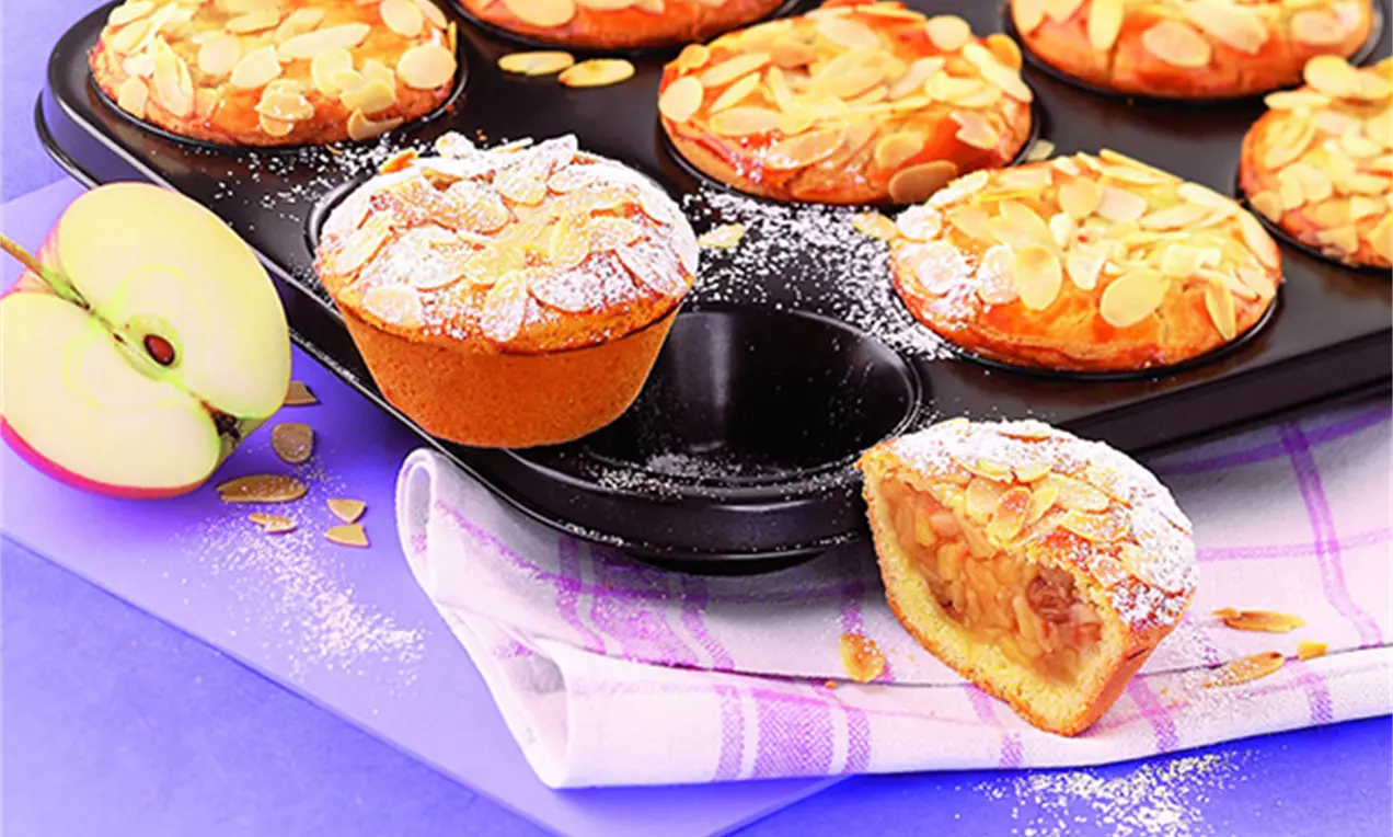 Goldbraune, runde Apfeltörtchen mit knusprigen Mandelblättchen und Puderzucker auf schwarzem Blech mit angeschnittener Füllung auf karierter Serviette