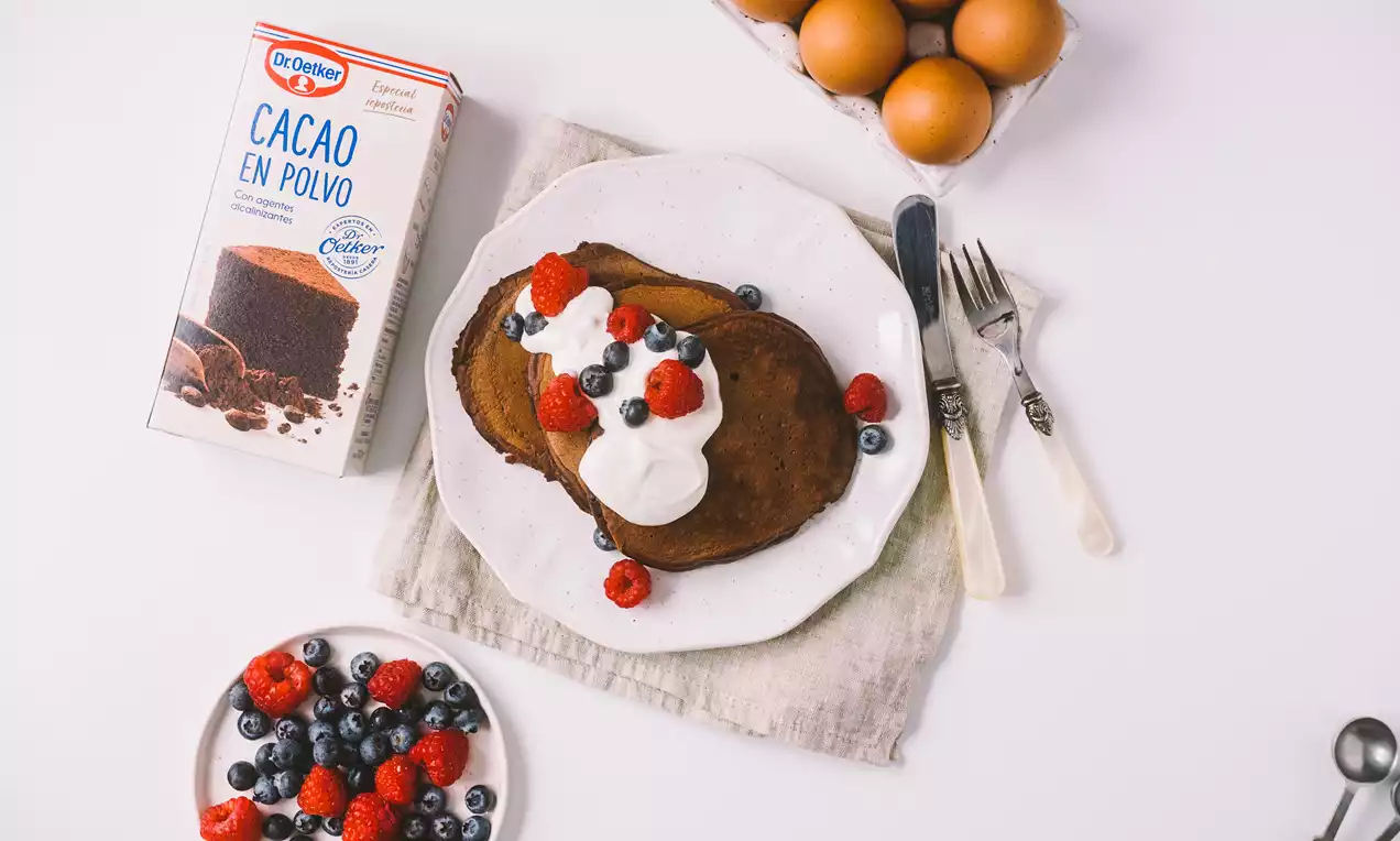 Tortitas de cacao de tono marrón oscuro con textura esponjosa, coronadas con crema blanca, frambuesas y arándanos sobre un plato blanco
