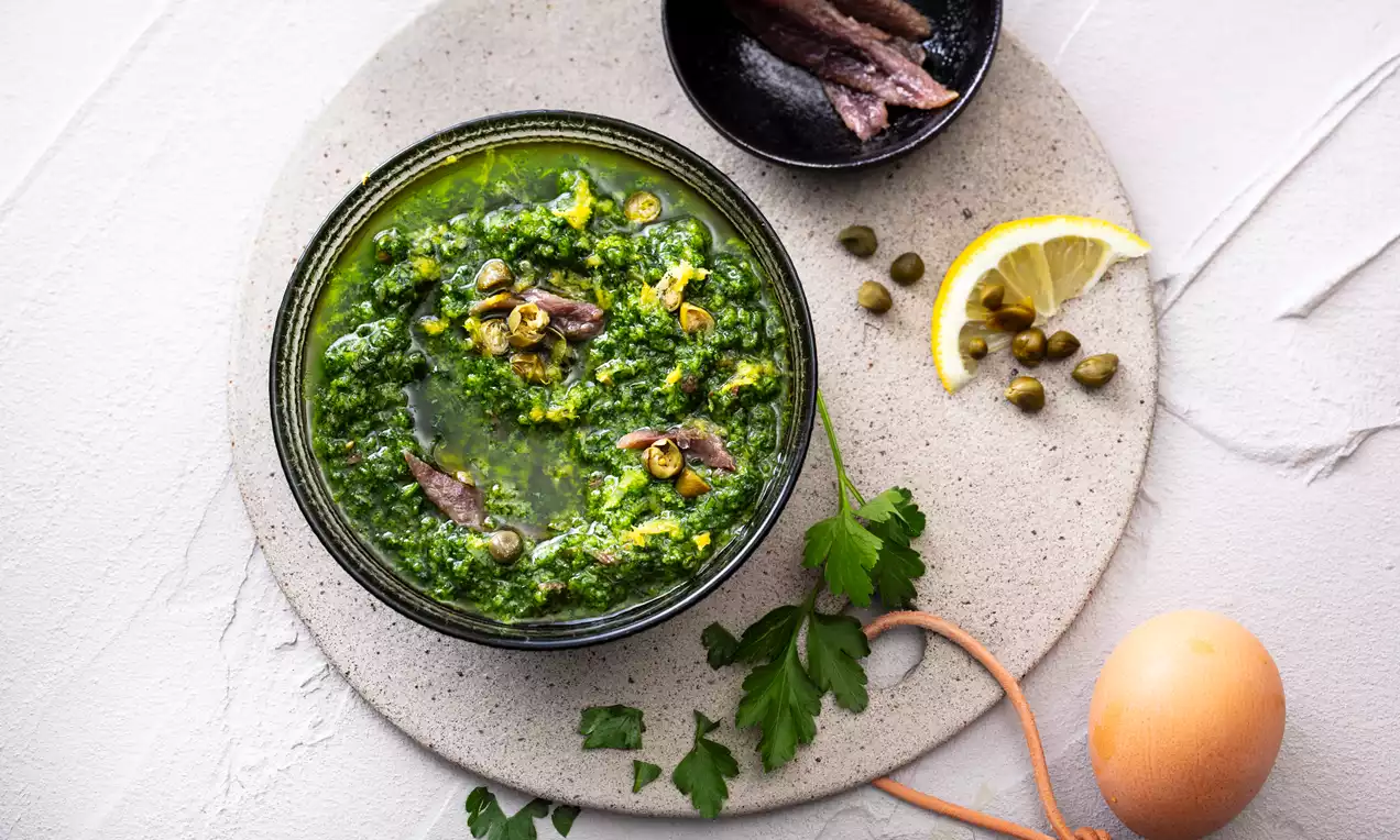 Salsa verde espesa y brillante con matices amarillos, coronada con anchoas y alcaparras, rodeada de limón, huevo y perejil sobre plato beige