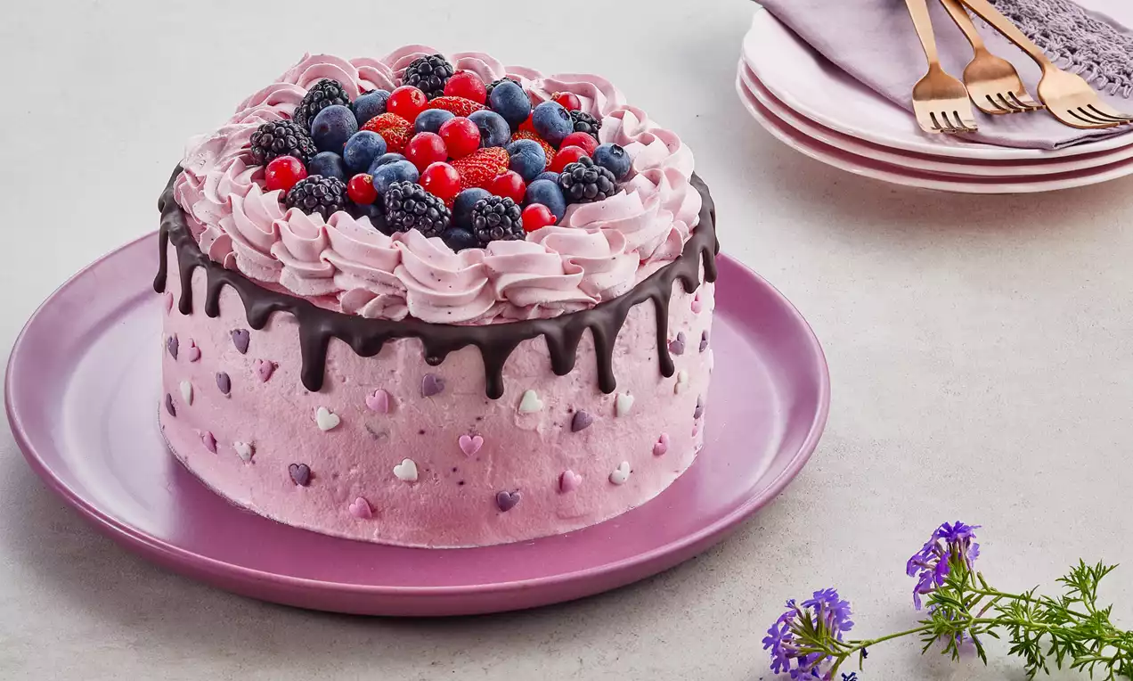 Pembe krema kaplı pasta, çikolata akıntıları, orman meyveli tepe süslemesi ve kalpli şeker detaylarıyla pembe tabakta