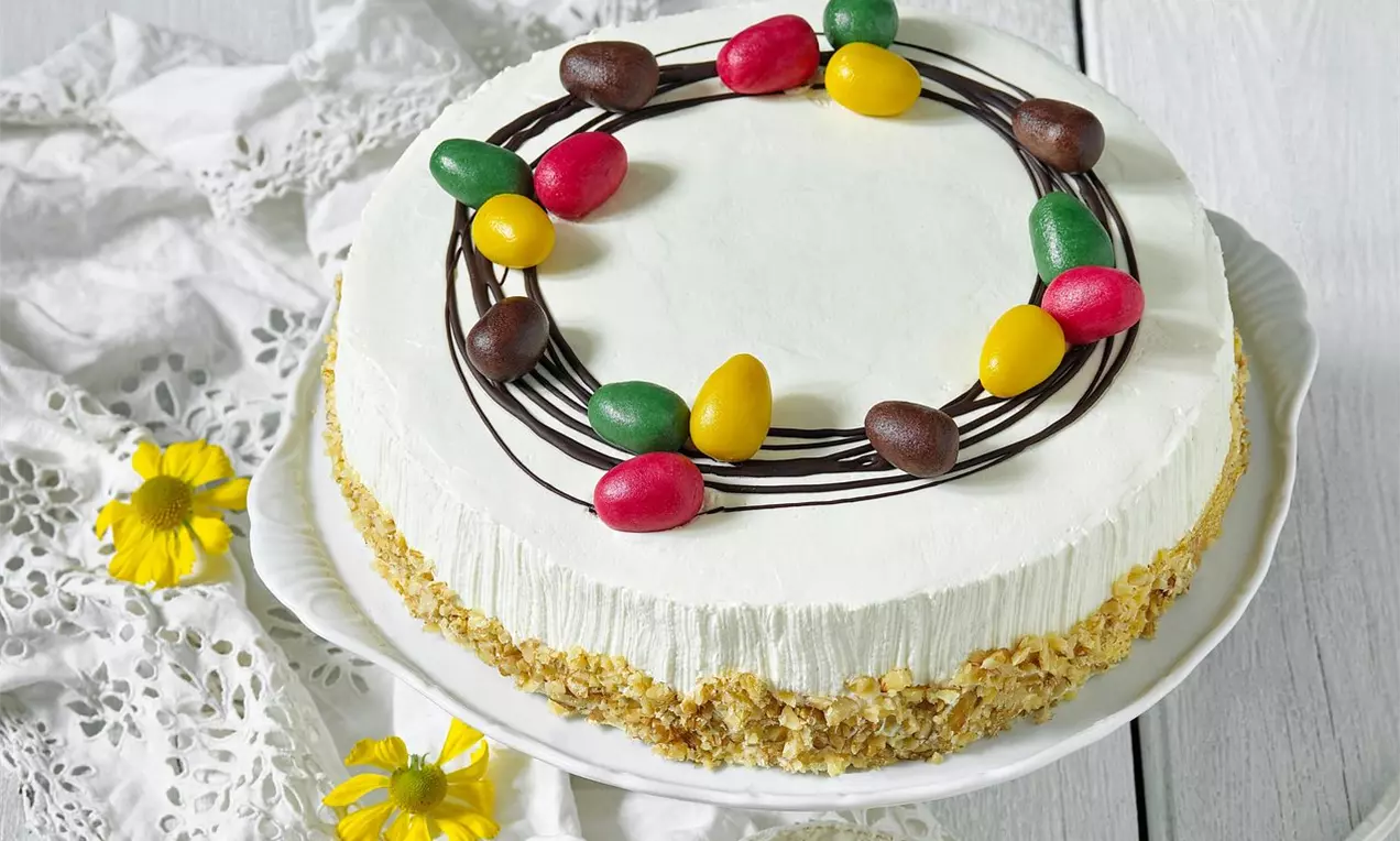 Velikonoční banánový dort s bílým hladkým krémem, zdobený barevnými cukrovými vajíčky na čokoládových liniích, sypaný oříšky na okraji
