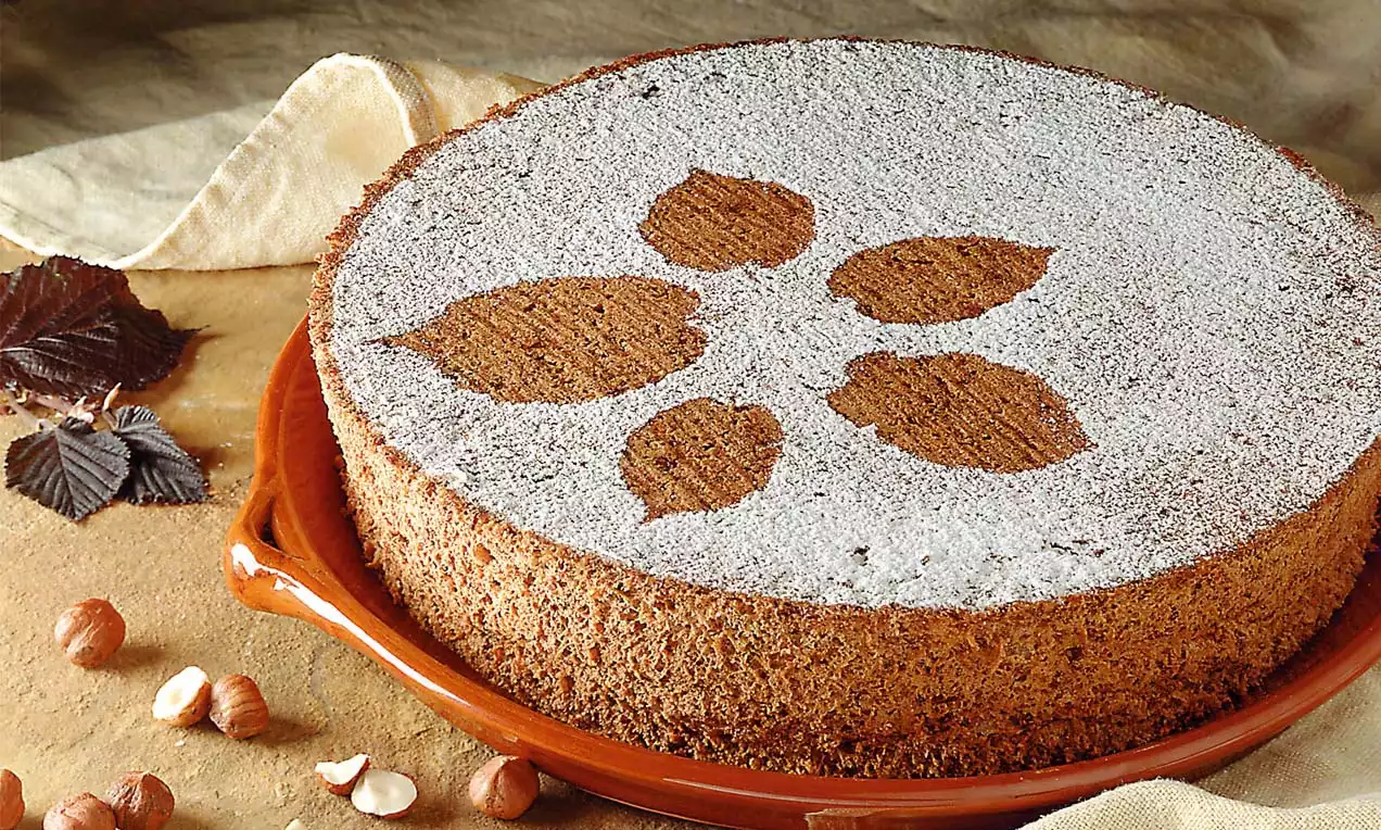 Runde Kartoffeltorte mit hellbrauner Kruste, bestäubt mit Puderzucker und einem Blattmuster verziert auf einer braunen Keramikplatte