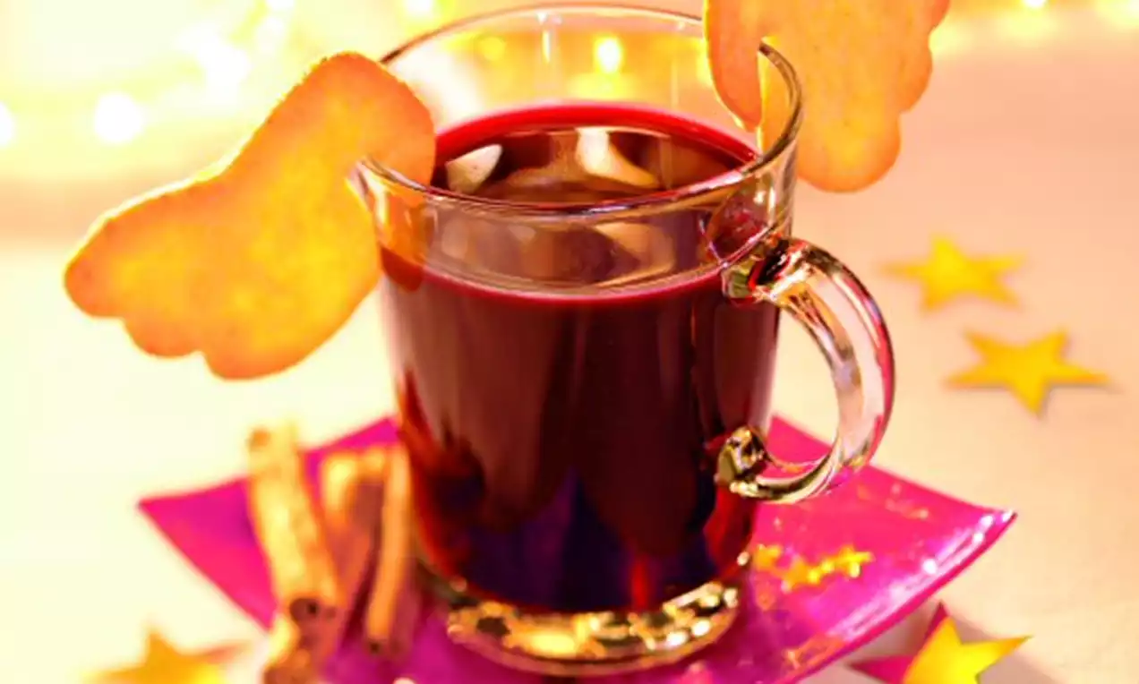 Ein Glas tiefroter Holunderpunsch mit zwei goldgelben Keksen in Engelsflügelform auf einer pinken Platte mit Zimtstangen und Stern-Deko