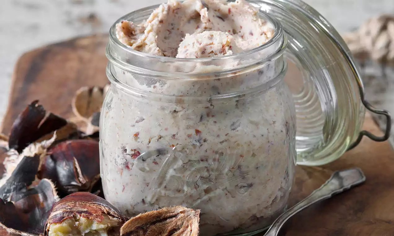 Kaštanové pyré světle hnědé barvy s viditelnými kousky, jemně krémové textury, naservírované v průhledné sklenici vedle louskaných kaštanů