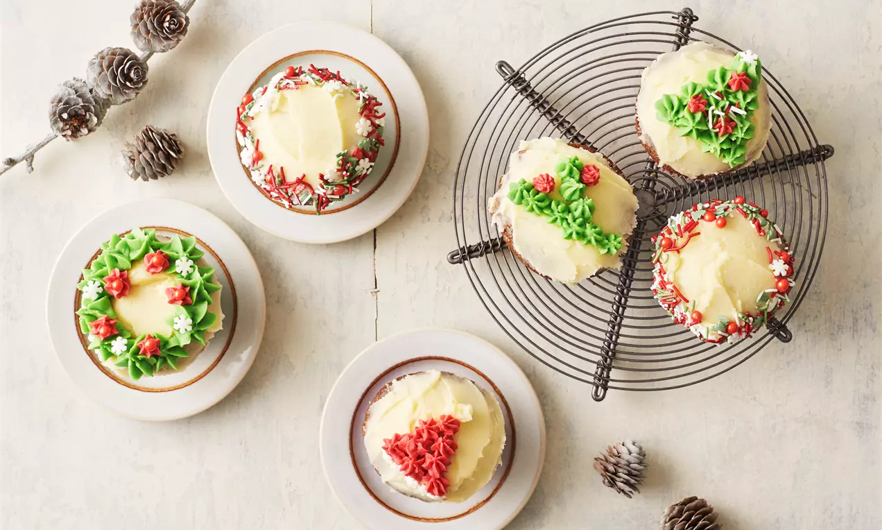 Jouluiset maustemuffinit samettisella voikreemillä, koristeltu punaisilla ja vihreillä sokerikuusilla ja kukilla, aseteltuina siivilän ja lautasten päälle