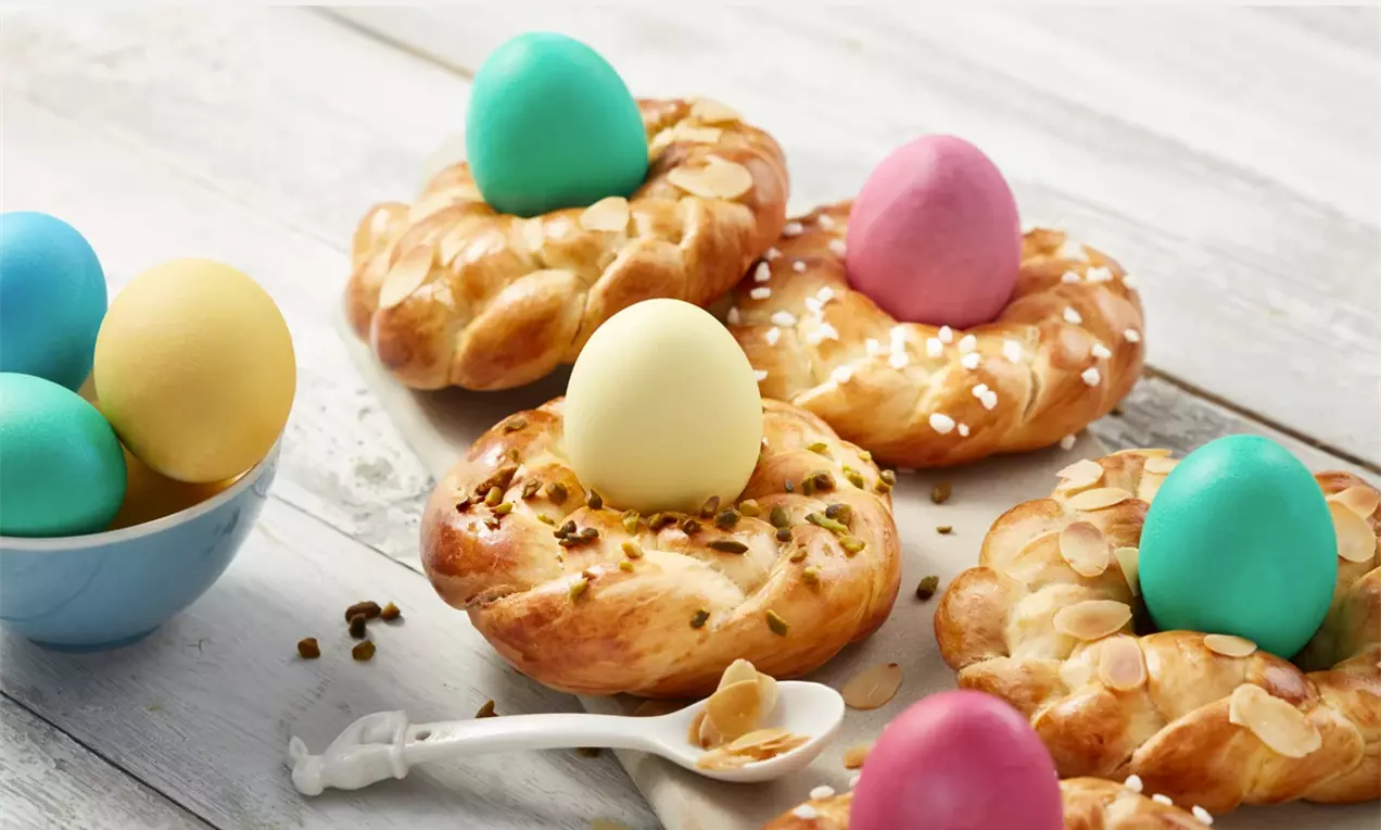 Златисто-кафяви козуначени венци с бляскава повърхност, поръсени с ядки и захар, украсени с яркоцветни яйца в центъра върху дървена поставка