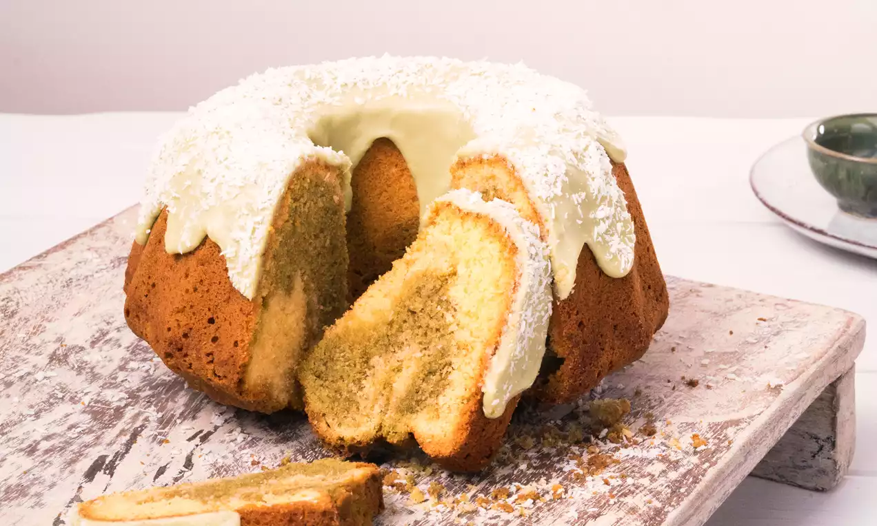 Kokos-Matcha-Marmorkuchen in goldbrauner Kranzform mit hellgrünem Zuckerguss und Kokosraspeln auf rustikalem Holzbrett angerichtet
