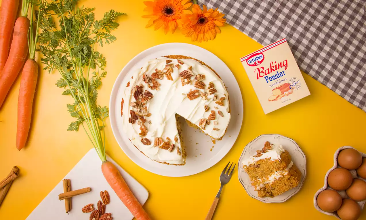 Tarta de zanahoria con cubierta de crema blanca esparcida, nueces troceadas, porción servida en plato pequeño sobre fondo amarillo