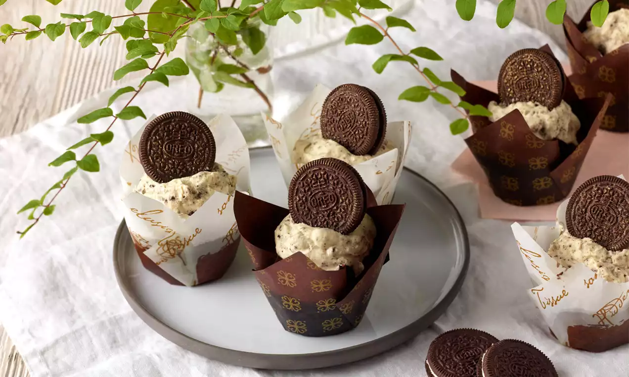 Oreo®-Cupcakes mit hellcremigem Topping und dunklem Oreo®-Keks, in braunen Papierförmchen auf grauem Teller neben grünen Zweigen und Keksen