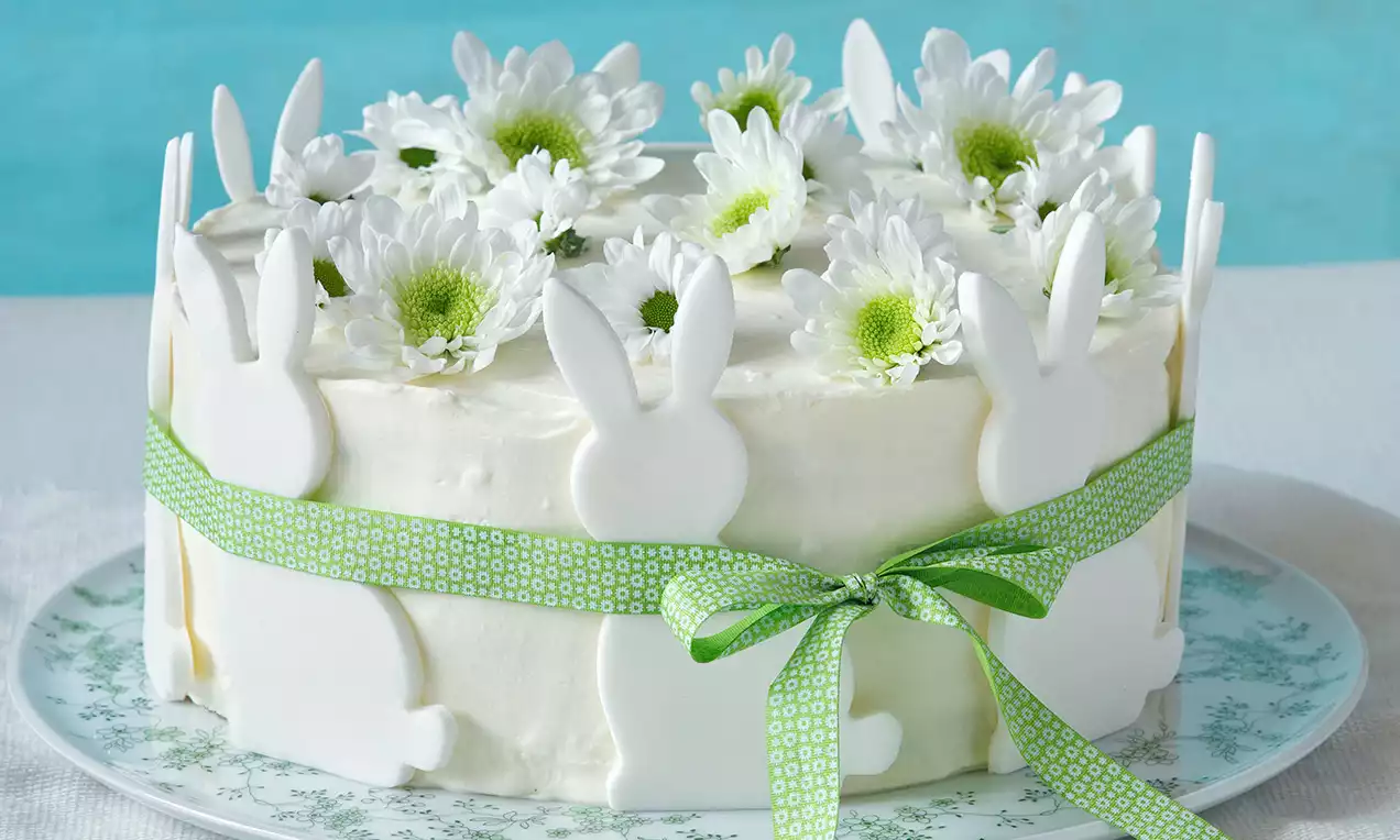 Velikonoční dort s hladkým bílým krémem, zdobený bílými sedmikráskami, obklopený cukrovými zajíčky a zelenou stuhou na vzorovaném talíři