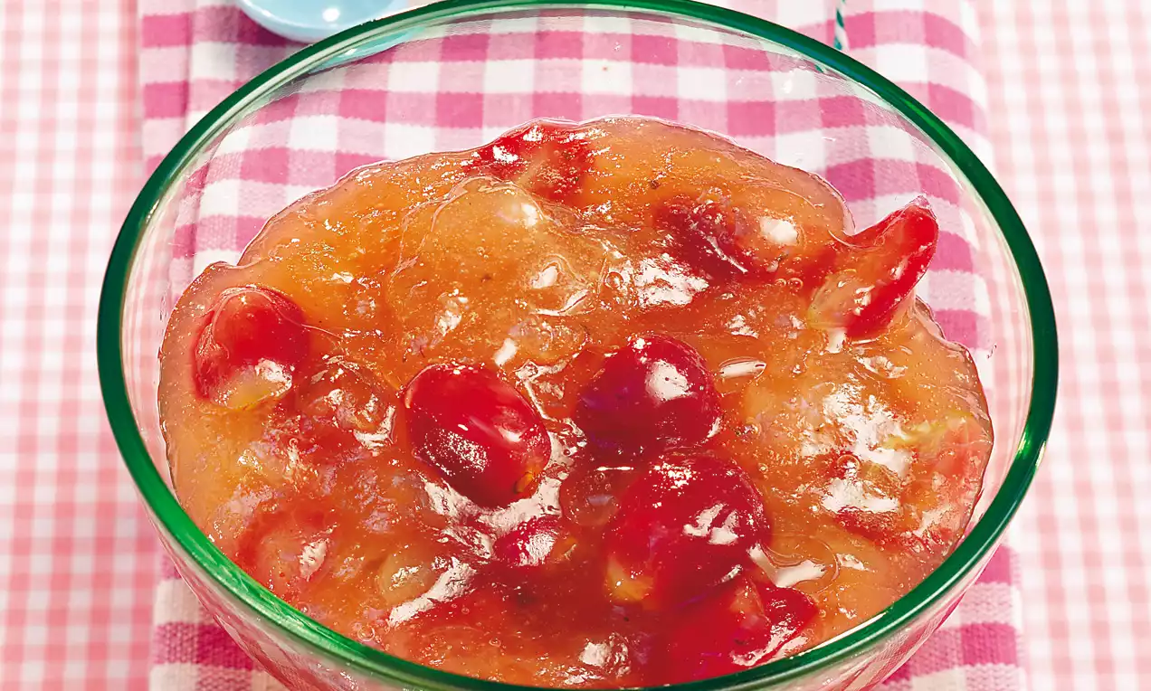 Confiture dorée translucide parsemée de morceaux de raisin rouge brillant dans un bol en verre sur une nappe à carreaux roses et blancs