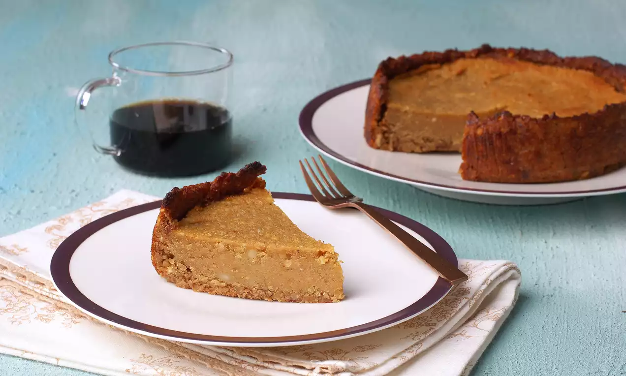 Kahverengi bisküvi tabanlı dilim limonlu vegan cheesecake, kıvamlı kreması ile beyaz tabakta bakır renkli çatal eşliğinde sunulmuş