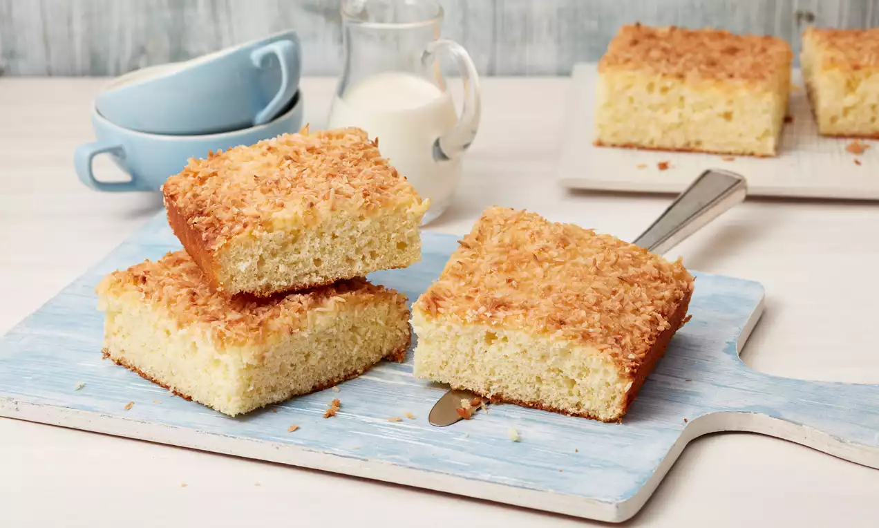 Zwei goldene, fluffige Kuchenstücke mit knuspriger Kokoskruste auf hellblauem Holzbrett, im Hintergrund Milch und weitere Kuchenstücke