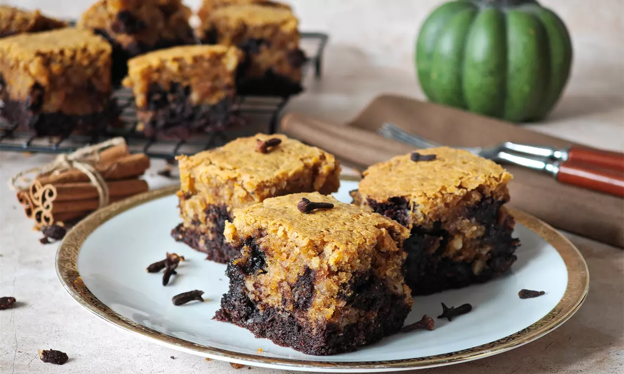 Квадратни парчета брауни с тиква, горен слой в топъл оранжев оттенък и долен наситено шоколадов, сервирани върху бяла чиния с канелени пръчици