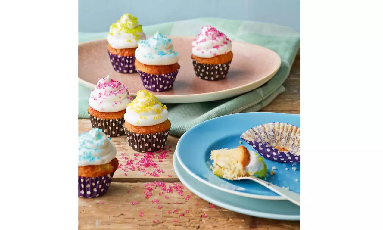 Pienet cupcake-leivokset tummissa paperivuoissa, kuohkea valkoinen kuorrute koristeltu kirkkailla sokerirakeilla, esillä lautasilla