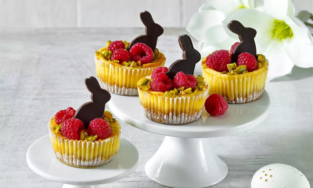 Mini cheesecakes v svetlých košíčkoch, ozdobené čokoládovými zajacmi, malinami a zelenými pistáciami na bielych podnosoch s veľkonočnou dekoráciou