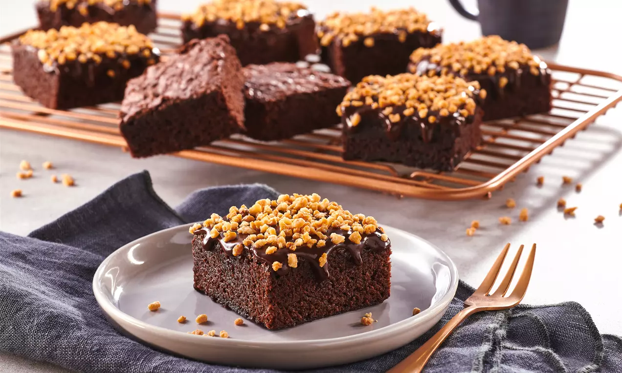 Saftiger dunkler Schokoladenbrownie mit glänzender Schokoladenglasur und goldbraunem Mandelkrokant auf einem grauen Teller mit Stoffserviette