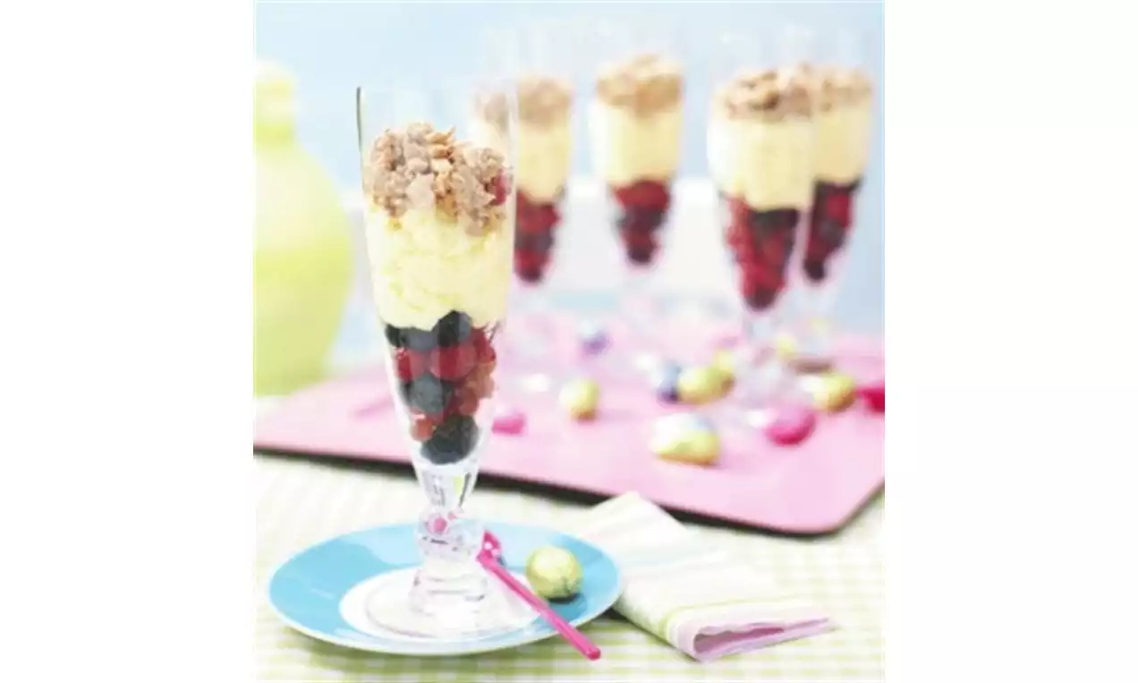 Hoge glazen gevuld met lagen donkerrode en paarse bessen, lichtgele mousse en kruimeltopping, gepresenteerd op een roze ondergrond met pastelpaaseitjes