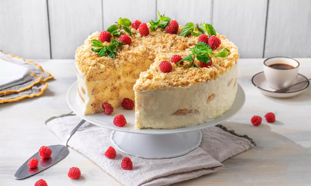 Кремообразна торта с лек ванилов оттенък, поръсена с хрупкави трохи и гарнирана с свежи малини и зелени листа върху бяла стойка