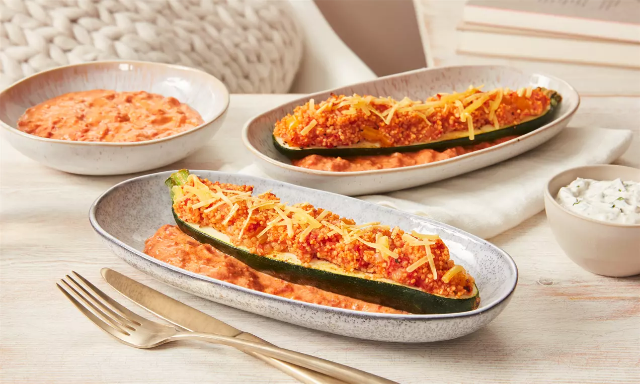 Halbierte grüne Zucchini, gefüllt mit rotgoldenem Belag und gelbem Käse, serviert auf ovalem Teller mit cremiger Beilage und Kräuterdip