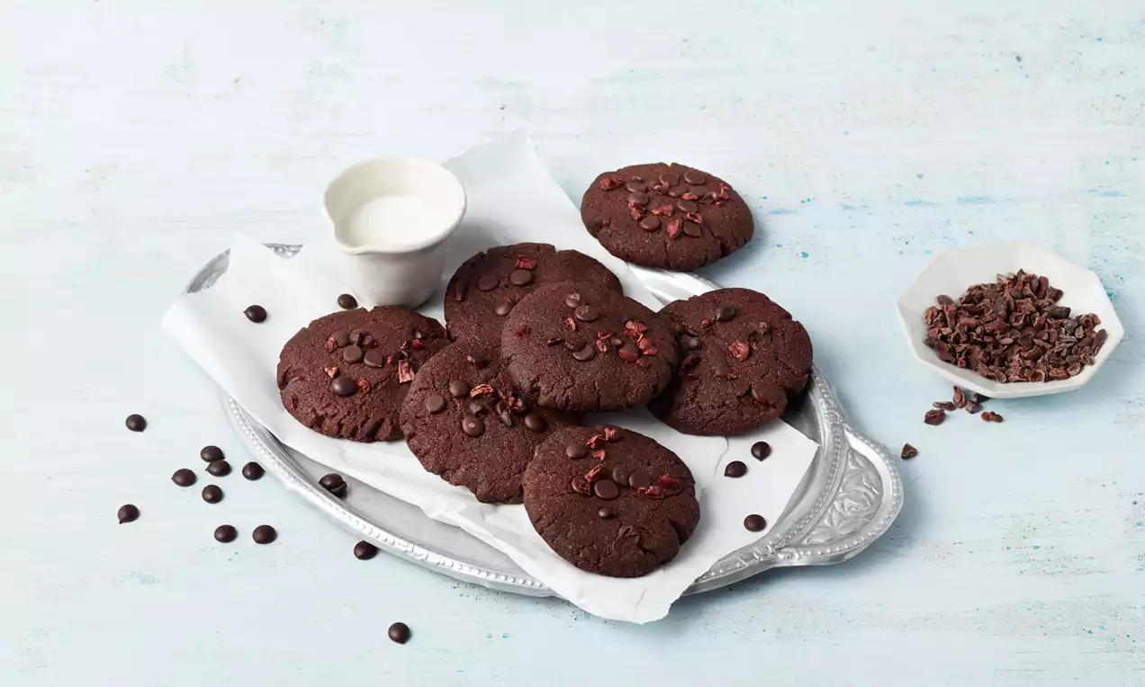 Runde, dunkelbraune Schokokekse mit knuspriger Textur und Schokostückchen auf Backpapier auf silberner Platte mit Milch und Kakaonibs