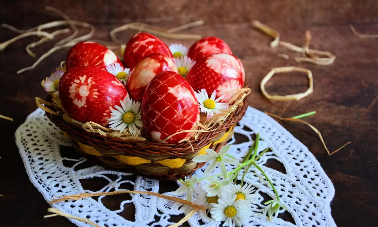 Crvena uskršnja jaja sjajne površine sa belim šarama leže u pletenoj korpici ukrašenoj belim tratinčicama na heklanom podmetaču