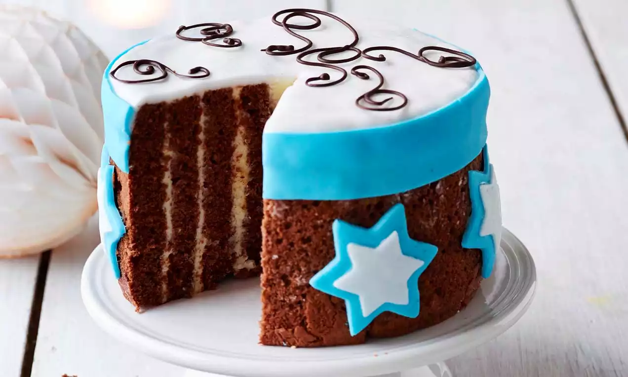 Lagkake med sjokoladebunn og lyse lag fyll, pyntet med hvit glasur, blå bånd, stjerner og sjokoladesnirkler på hvitt kakefat
