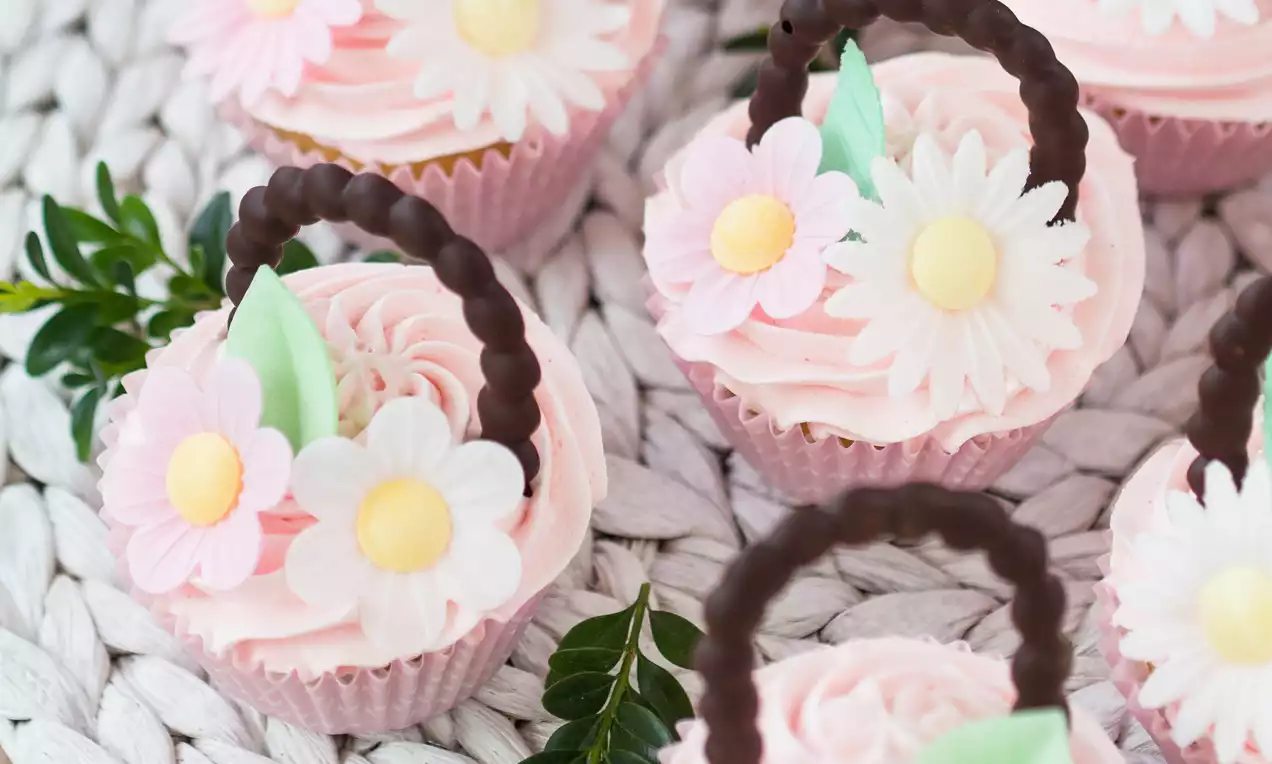 Babeczki w różowych papilotkach ze słodkimi uchwytami z czekolady, dekorowane różowymi i białymi kwiatkami oraz zielonymi listkami na plecionym tle