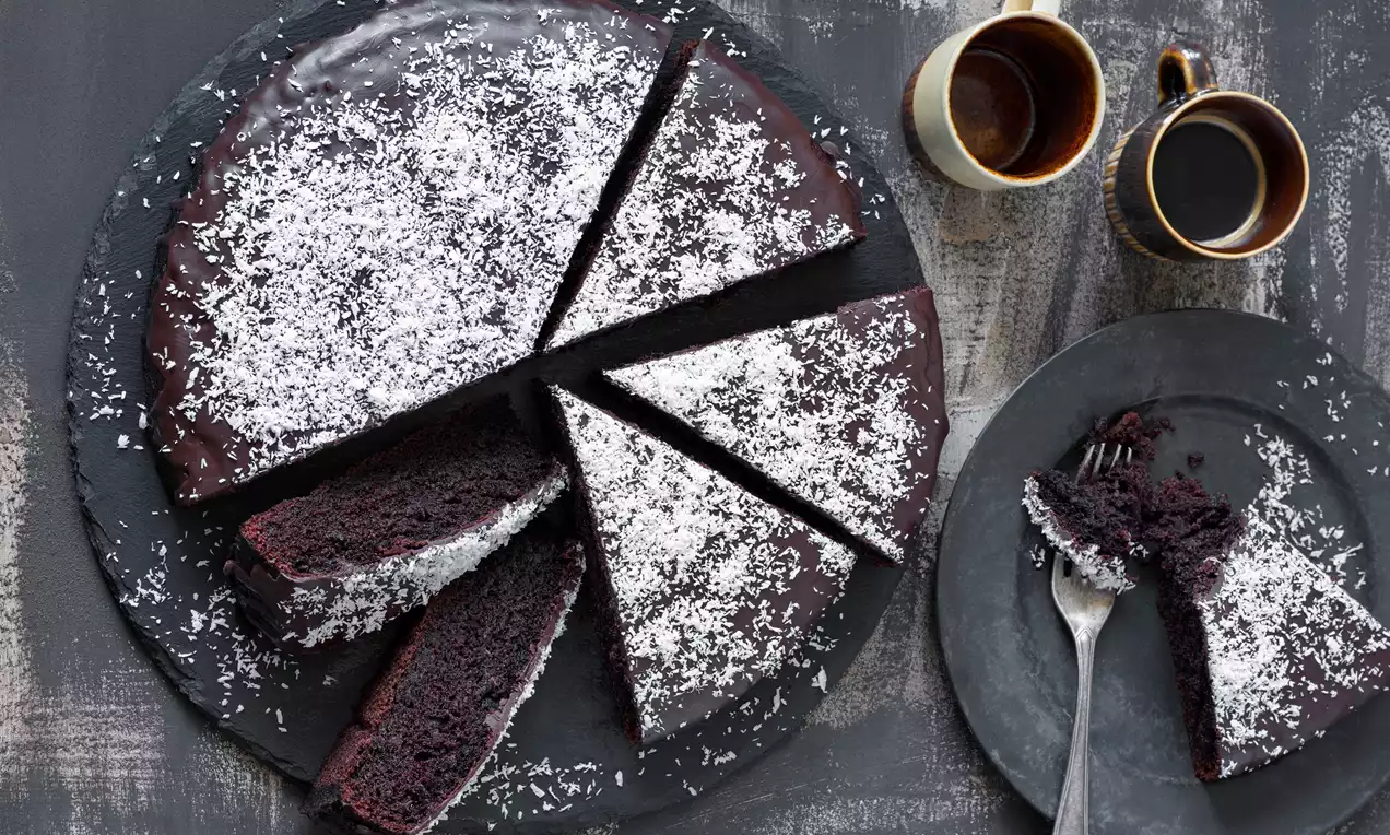 Tmavá kruhová buchta s lesklou čokoládovou polevou, posypaná strouhaným kokosem, nakrájená na trojúhelníkové kousky na černém talíři