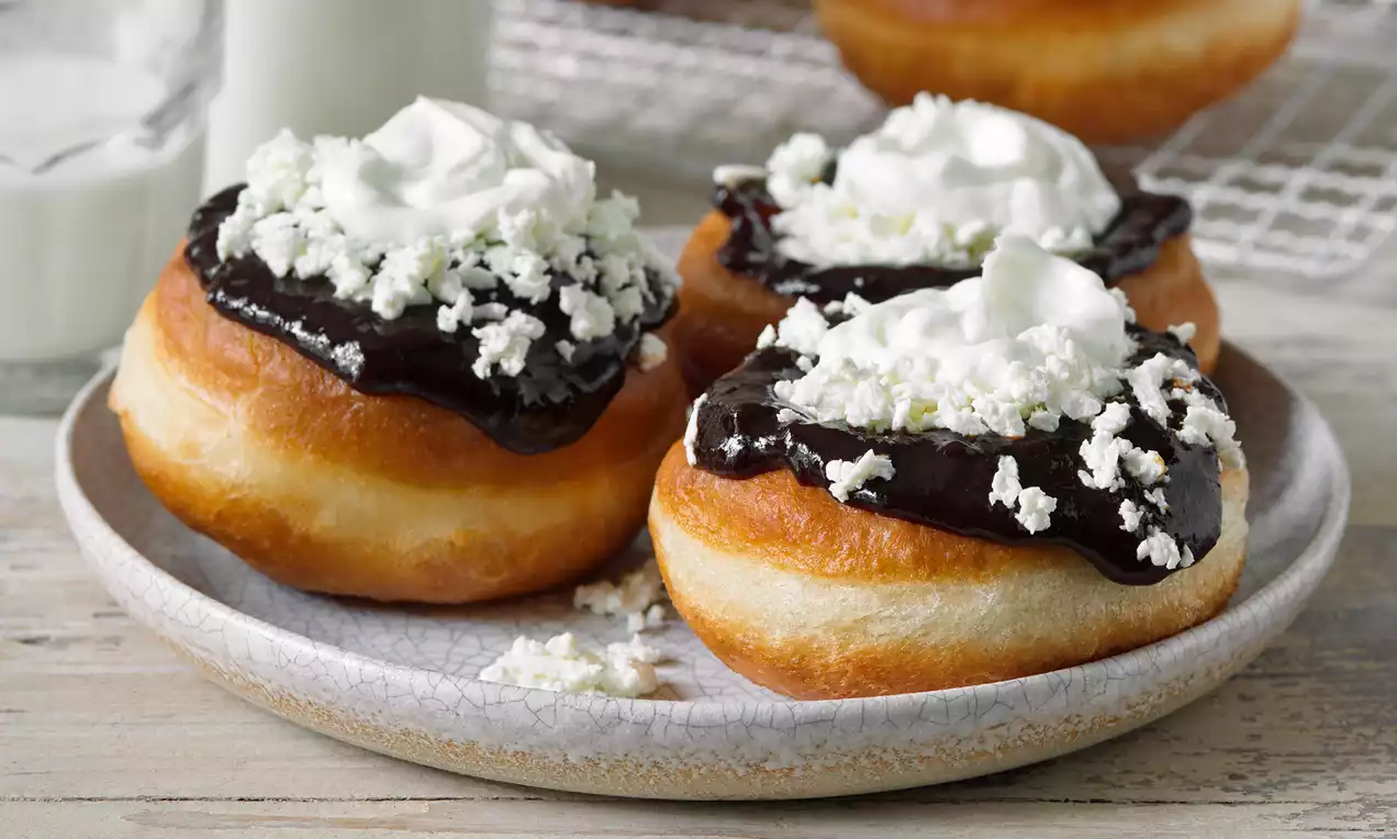 Zlatavé kulaté vdolky s lesklou vrstvou tmavé čokoládové polevy, zdobené bílou šlehačkou a drobeným sýrem na rustikálním talíři