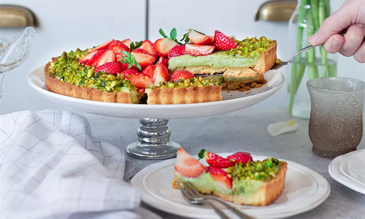 Тарт с златист хрупкав блат, кремообразен зелен шамфъстъчен пълнеж, поръсен с натрошен шамфъстък и свежи червени ягоди върху бяла стойка