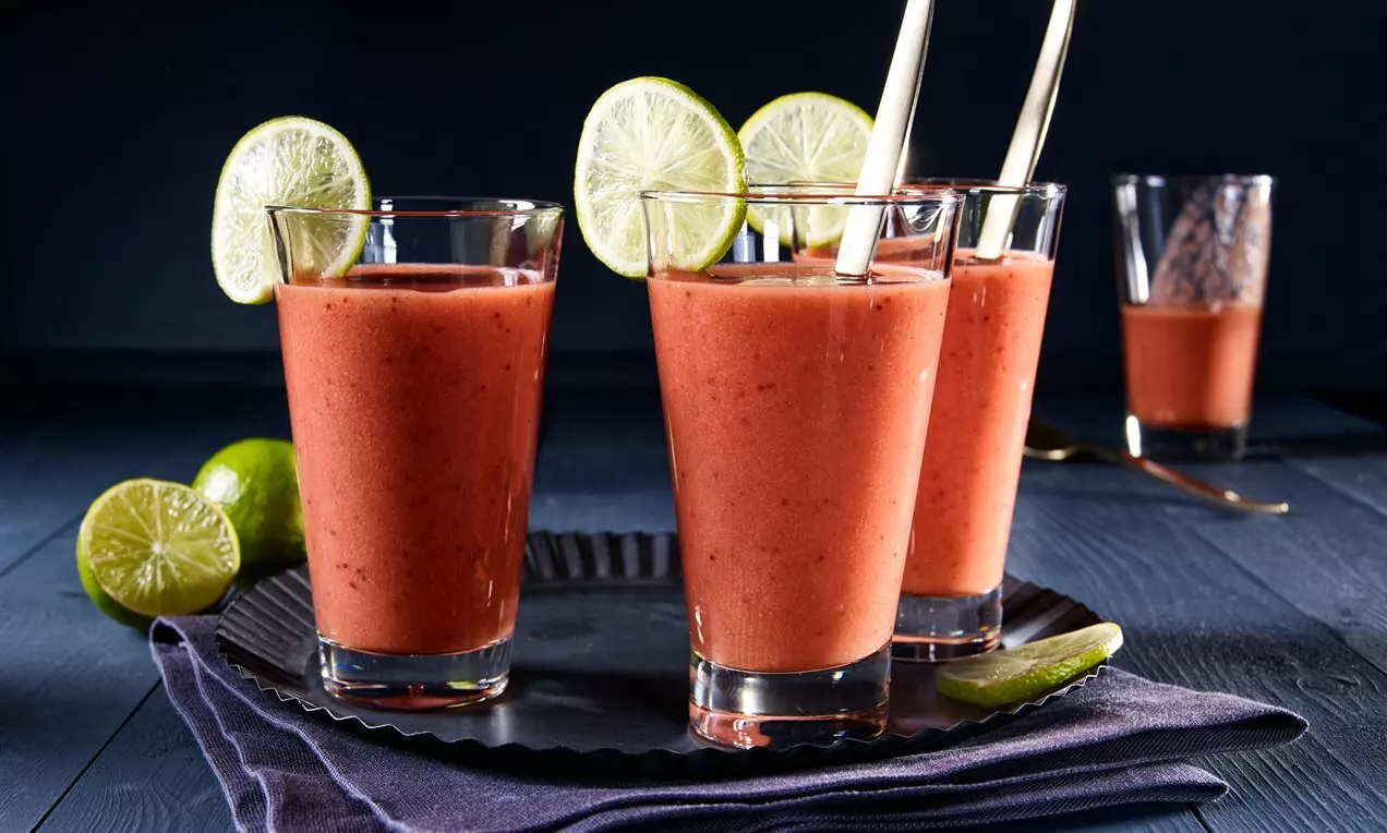 Четири високи стъклени чаши с гъсто розово смути, украсени с резенчета лайм, върху тъмносин плат и черен метален поднос