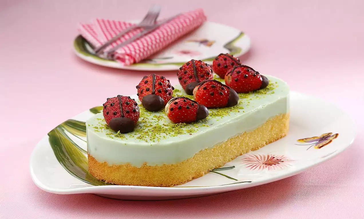 Pasta altın sarısı tabanı, yeşil kreması, çikolata kaplı çilek süslemesi ve çiçek desenli tabak üzerinde sunulmuş