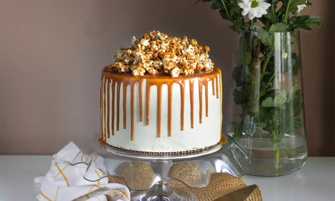 Висока кръгла торта с бяла глазура, карамелени капки и купчина златисти пуканки отгоре, поднесена на стъклена стойка