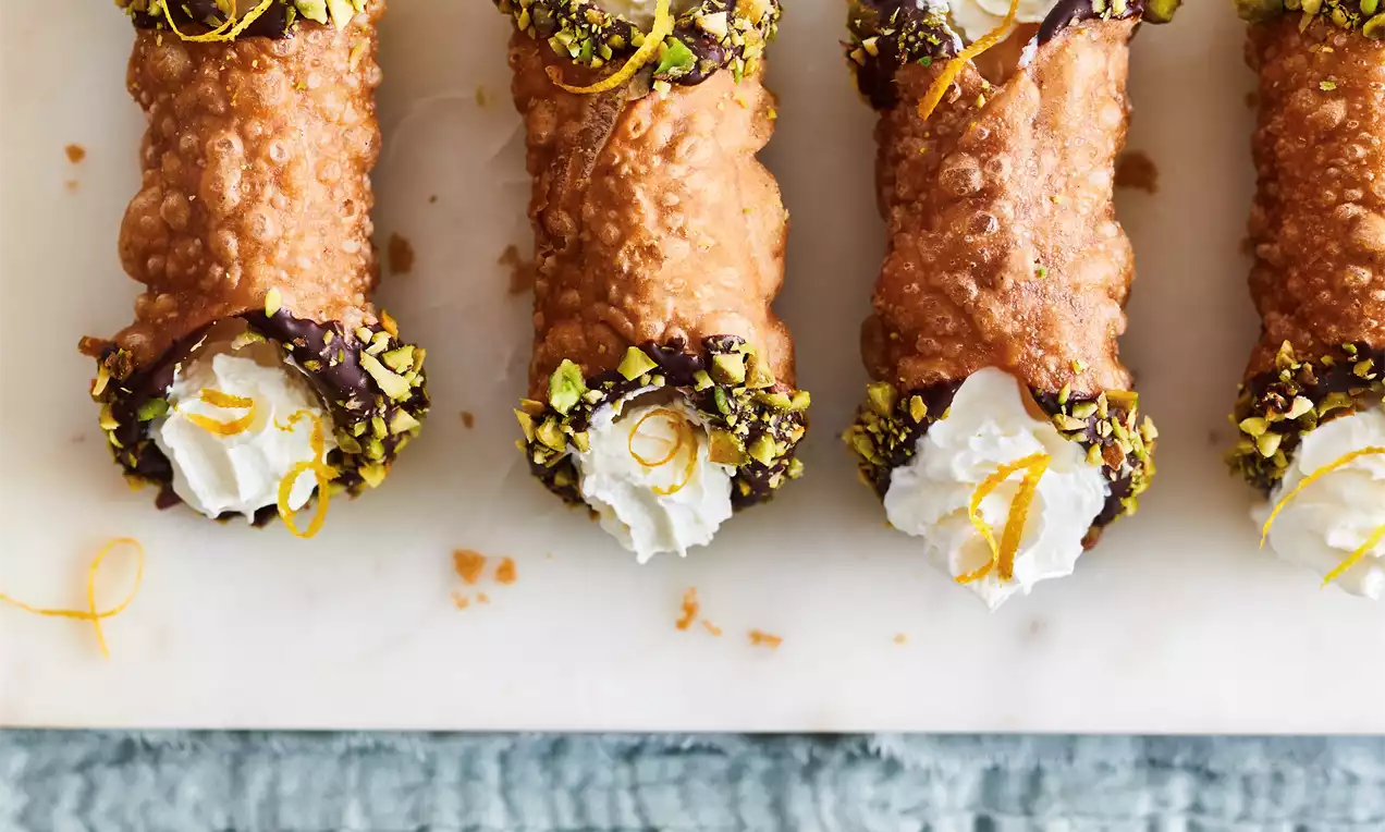 Golden-brown cannoli shells with textured ridges, filled with white cream, dipped in chocolate and pistachios, garnished with lemon zest