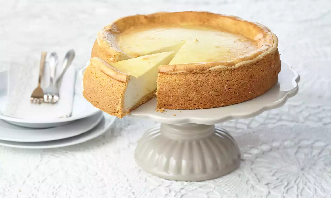Goldbrauner Käsekuchen mit glatter, heller Oberfläche und einem Stück herausgeschnitten auf weißer Tortenplatte mit Teller und Besteck im Hintergrund