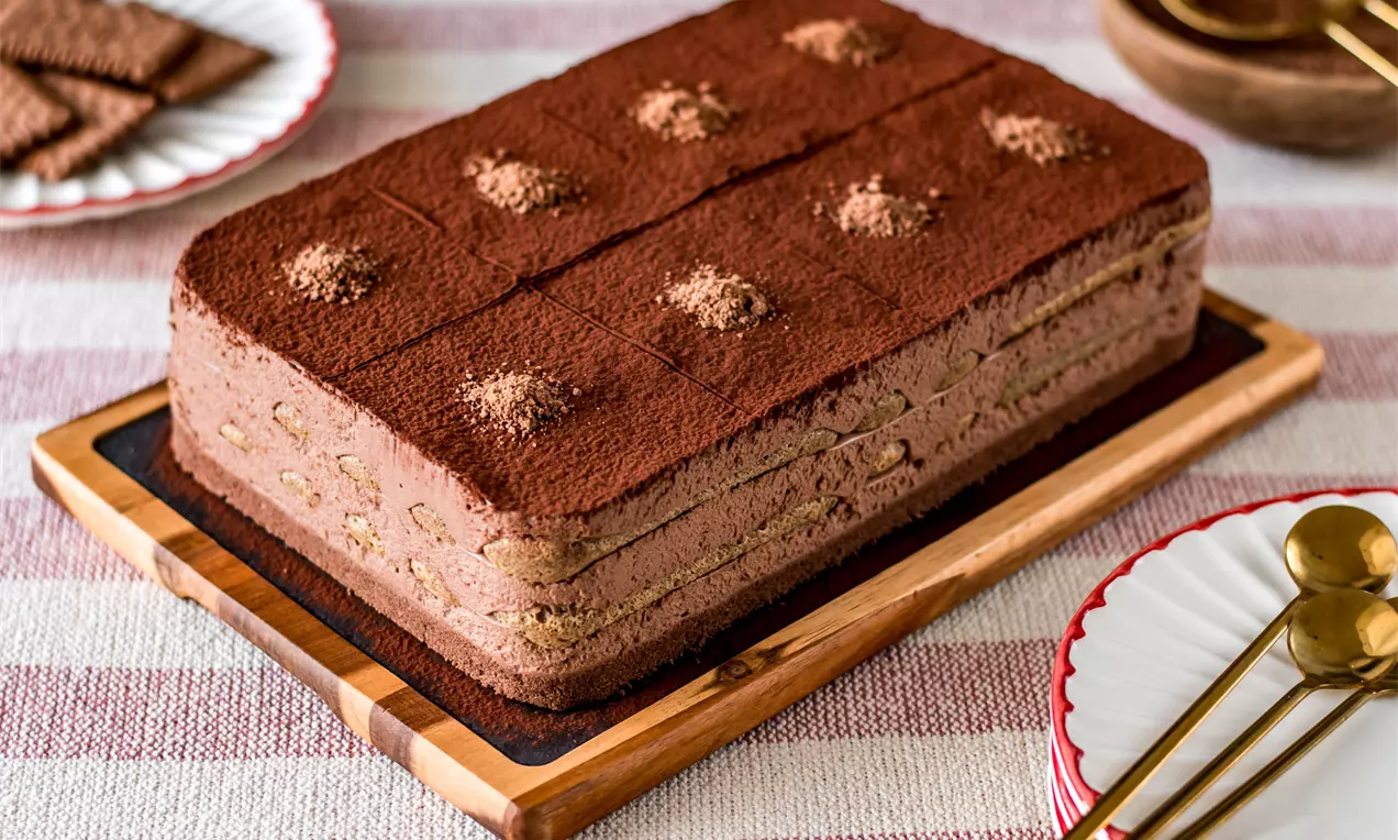Правоъгълна торта с кафяв бисквитен основен слой и крем с текстура на пухкаво мляко с какао, украсена с какаов прах върху дървена поставка