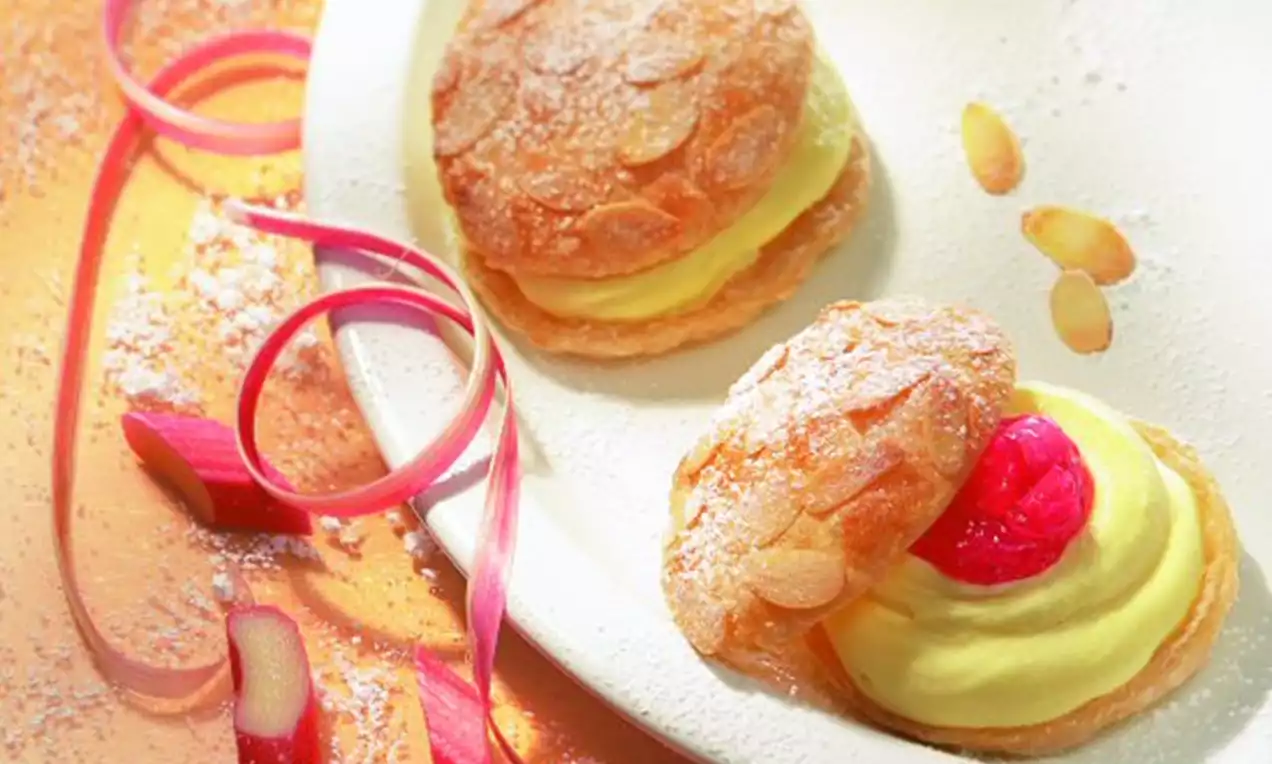 Goldbraune Blätterteighälften mit gerösteten Mandelblättchen, gelber Creme und roter Rhabarberfüllung auf weißem Teller mit Rhabarberstreifen