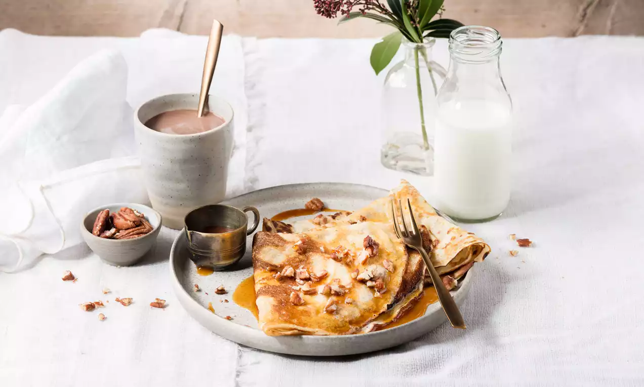 Goudbruine pannenkoeken licht opgevouwen met glanzende karamelsaus en bruine stukjes noten op een grijs bord met een gouden vork