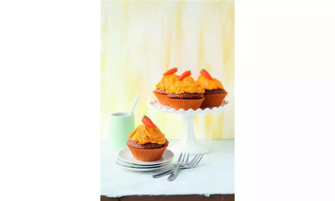 Gulrotcupcakes med fyldig oransje krem og små maripan-gulrøtter på kakestativ og tallerken, omgitt av gafler og lys bakgrunn