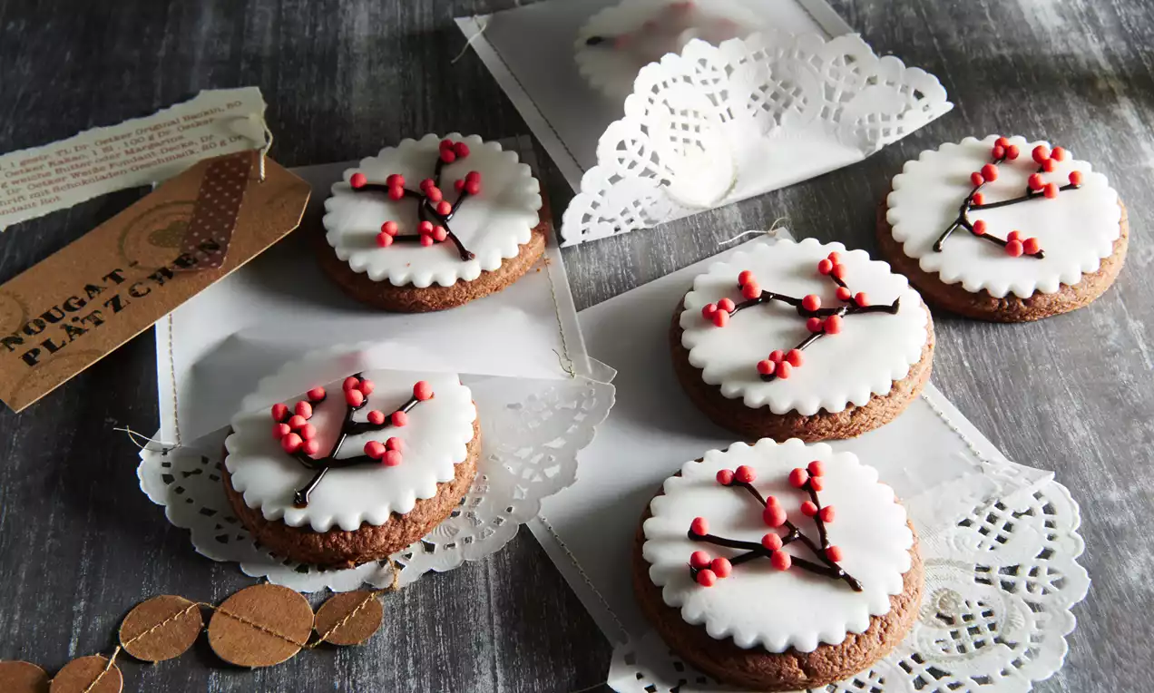 Okrugli nougat keksi tamnosmeđe podloge s bijelom glazurom i crvenim ukrasima u obliku bobica, posluženi na čipkastim papirima