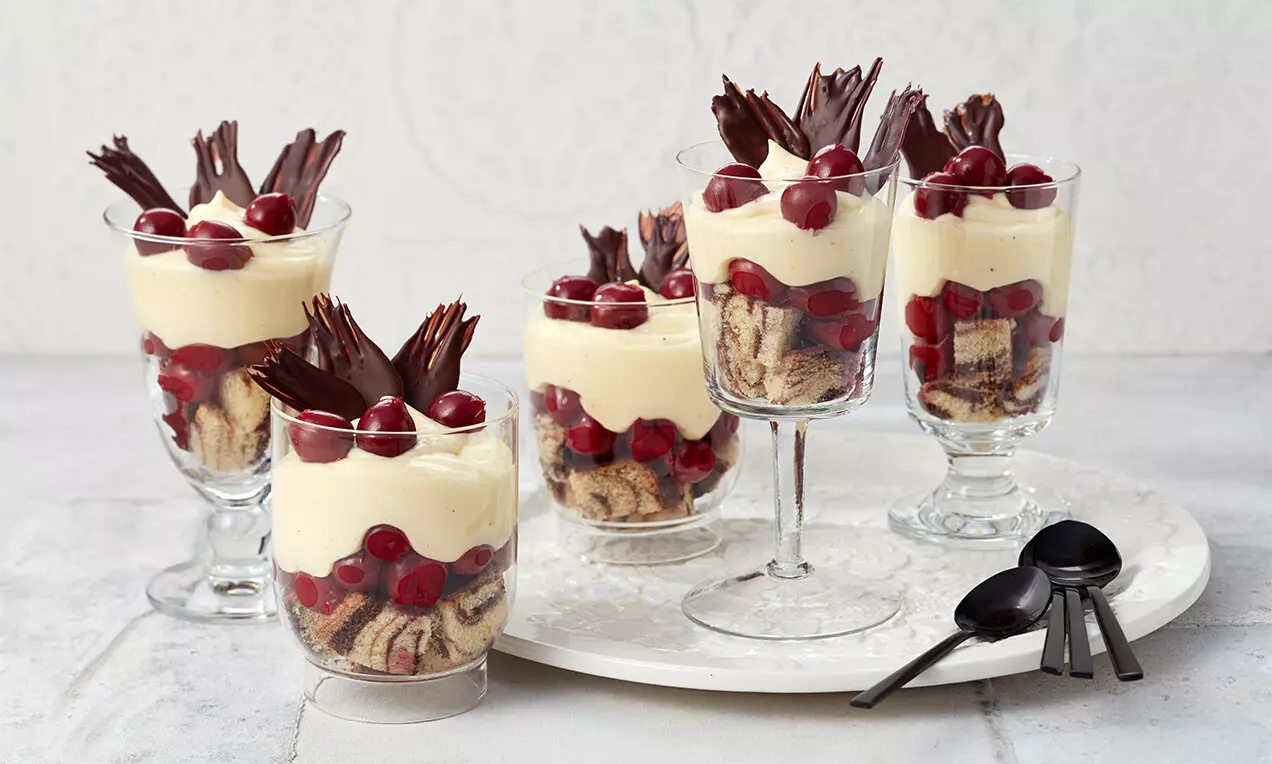 Слоеве от светъл крем, парчета шарен кекс и сочни червени череши в прозрачни чаши, украсени с тъмнокафяви шоколадови декорации